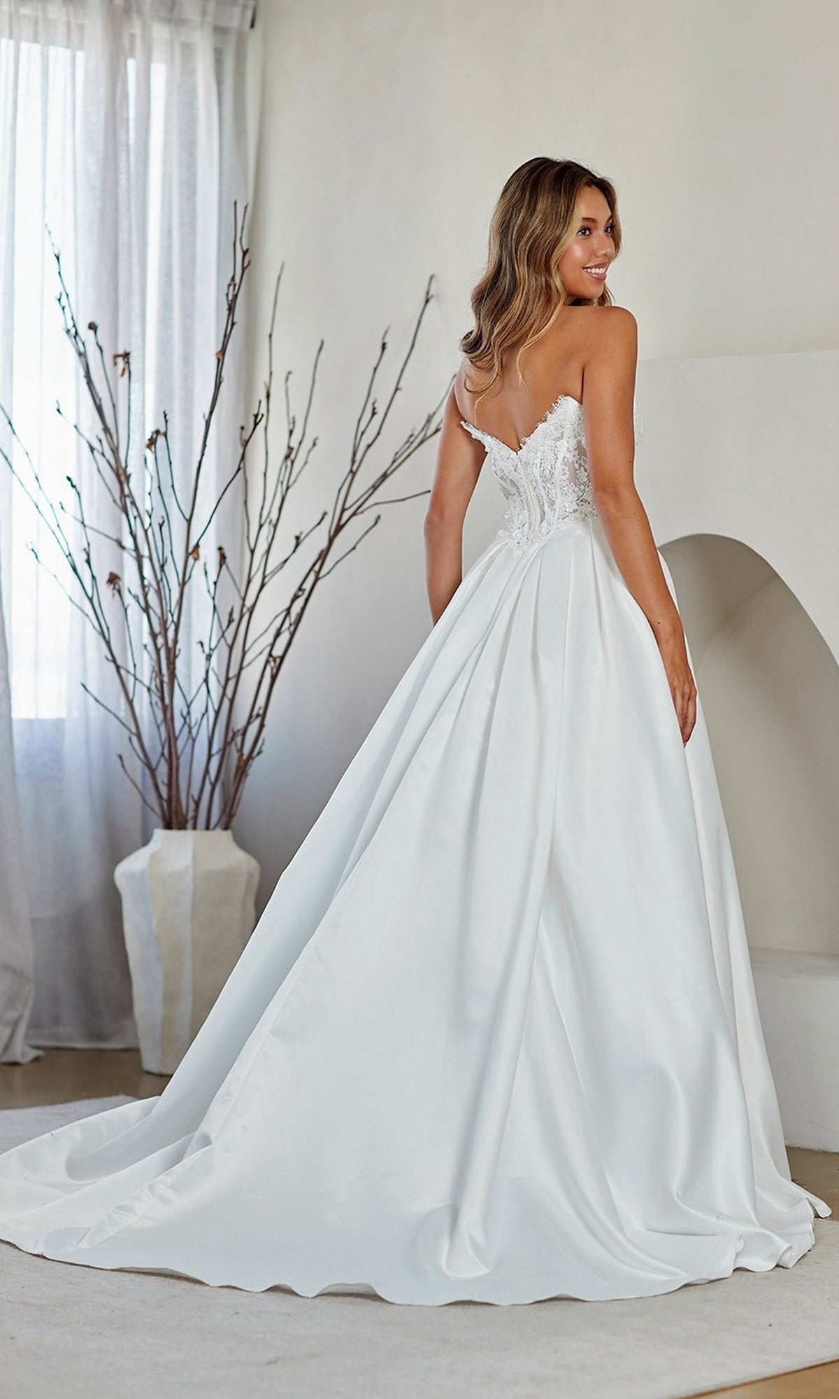 A woman stands indoors, turned to show the elegant back of the Amelia Couture 2089 Long Prom Dress—a white strapless ballgown with a lace corset bodice and long satin skirt.